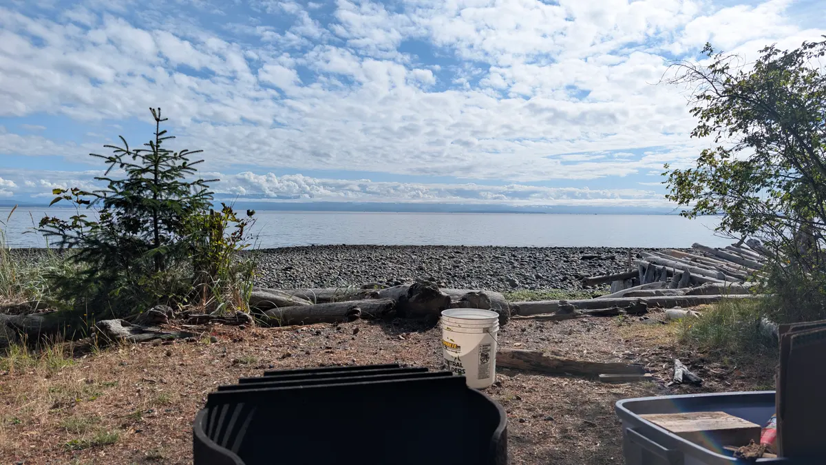A campsite fire pit above a driftwood beach, looking across the Strait