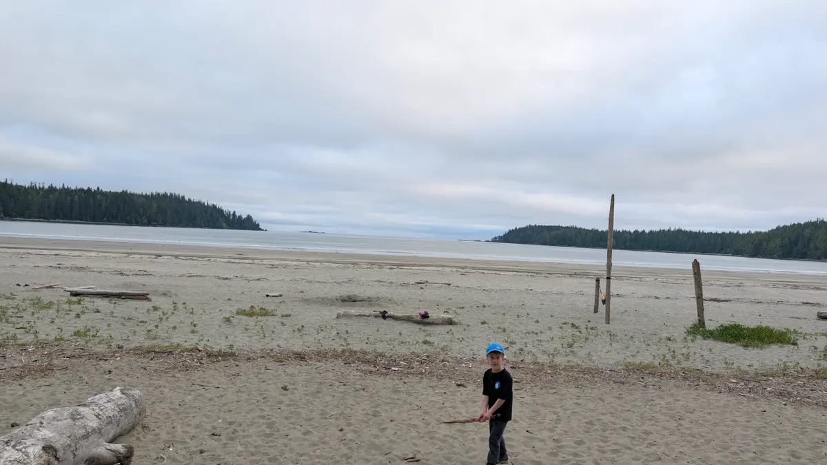 A child on a driftwood-strewn sand beach at Pachena Bay, with forested headlands across the water